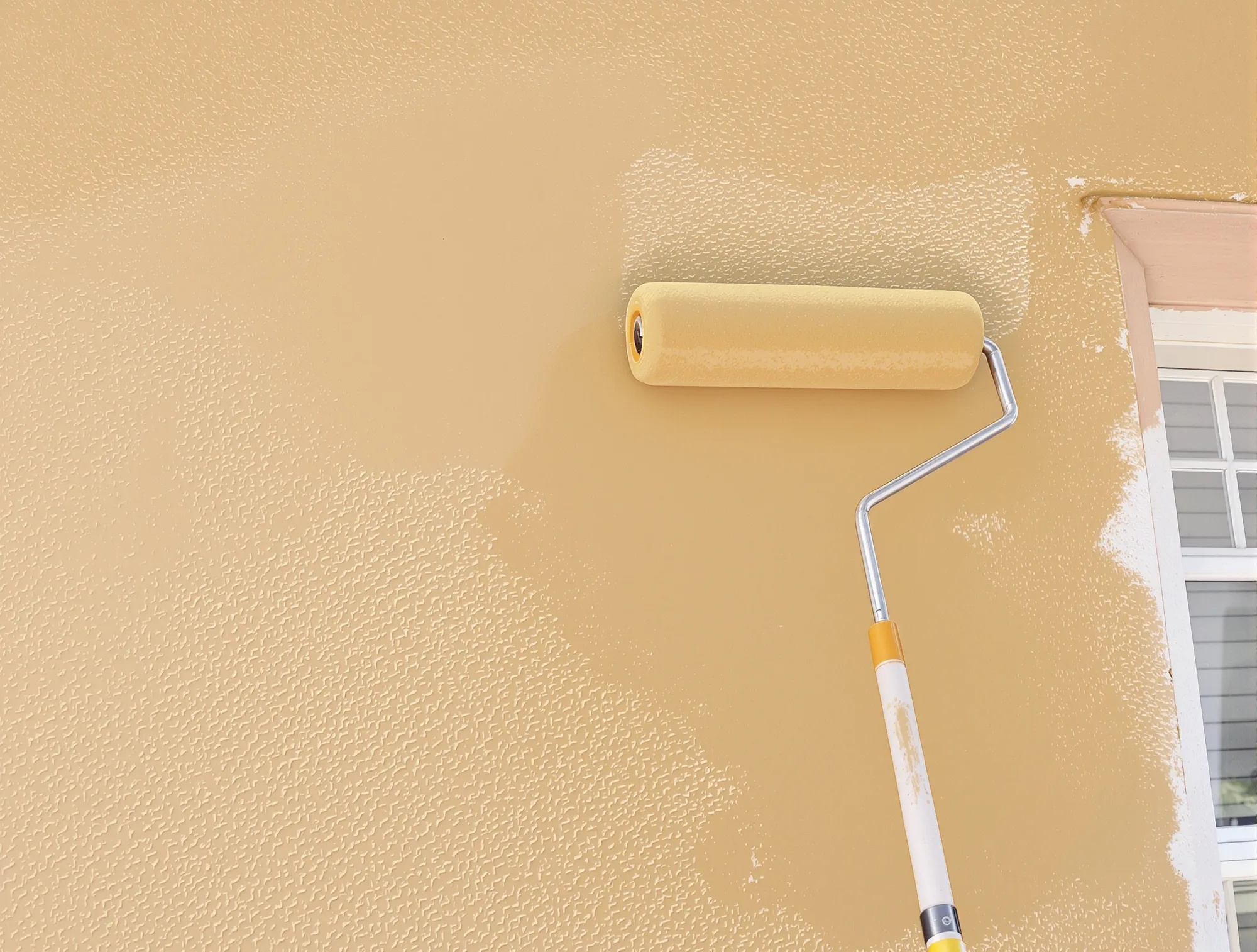 Roller on a pole back-rolling fresh tan paint into textured stucco