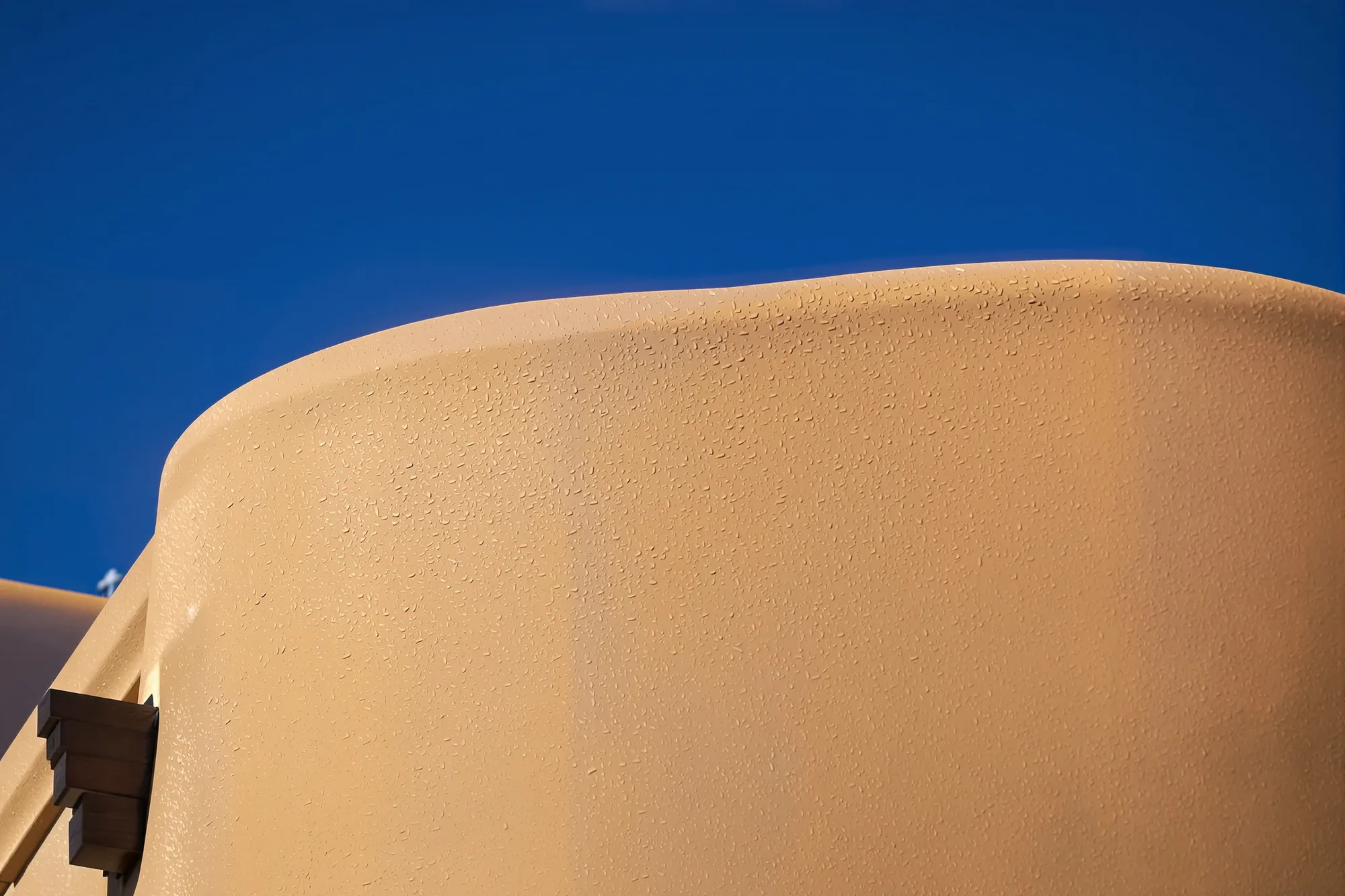Rounded adobe brown stucco parapet with dark viga ends and a metal canale against a blue sky